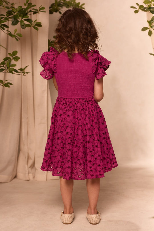 A young girl with curly brown hair stands facing away, wearing the Mini Hattie Dress in Magenta by Ivy City Co. The embroidered eyelet fabric, puffed sleeves, and delicate lace details create a charming scene, as she stands on a light floor surrounded by leafy branches and beige curtains—capturing the essence of a Mini & Me Easter dress moment.