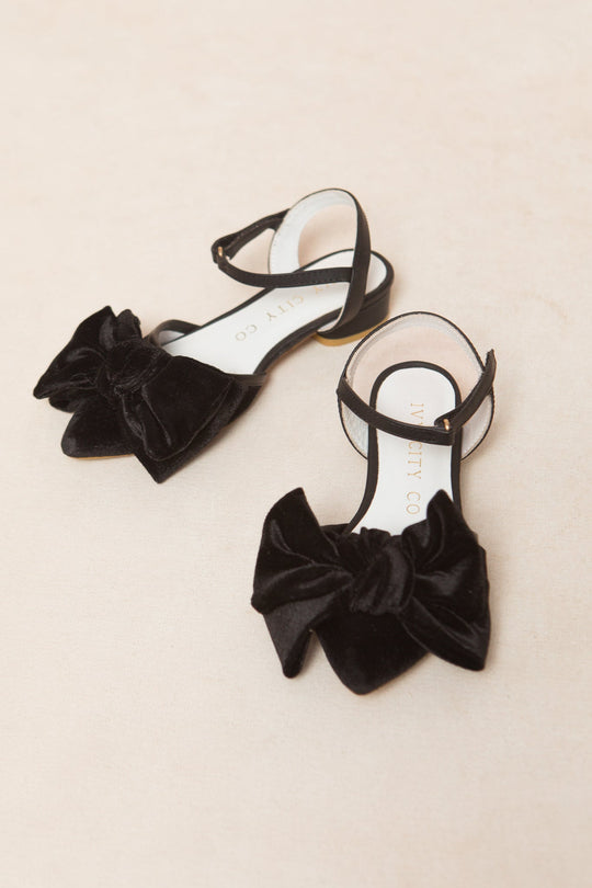 The Mini Eloise Bow Heel in Black by Ivy City Co features black velvet flats with ankle straps and large decorative bows on the toes, elegantly displayed on a light beige surface.