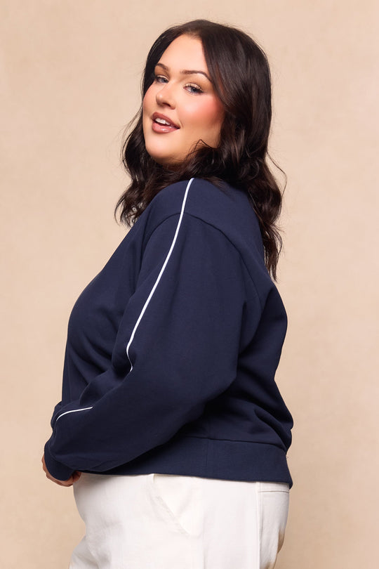 A woman with wavy dark hair, dressed in the Ivy City Co Kinsley Sweatshirt in Navy and white pants, poses against a beige background, smiling gently over her shoulder.