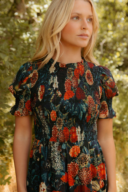 A woman with blonde hair stands outdoors amid greenery and sunlight, wearing the Cordelia Dress in Moody Floral by Ivy City Co and gazing calmly to the side.