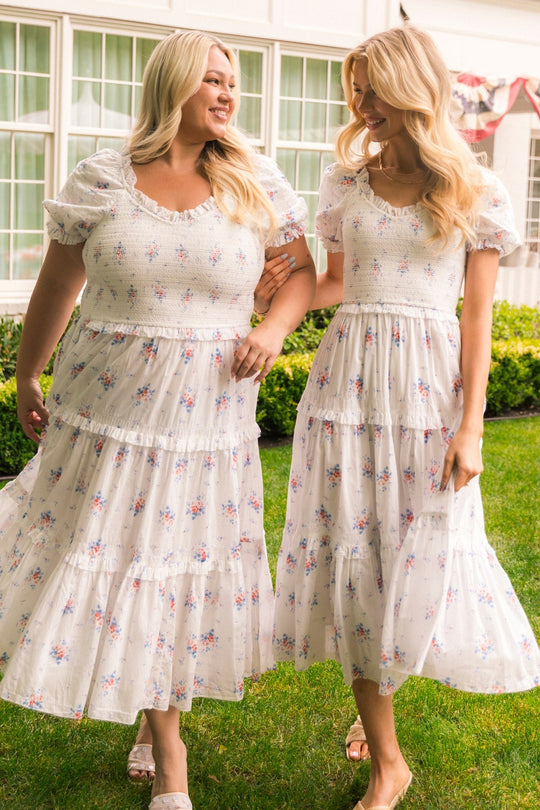 Two women in matching Emily Dress in Heritage Soft Ditsy Floral by Ivy City Co walk hand in hand on the grass, smiling at each other. They are outside near a white building with large windows and garden bushes. The nursing-friendly design of their Ivy City Co dresses adds both comfort and charm to this joyful moment.