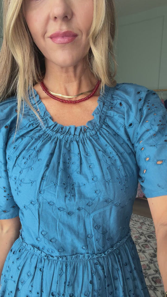 A woman wearing the Ivy City Co Etta Dress in Cobalt, featuring short sleeves, and a red beaded necklace stands indoors facing the camera. Only her upper body is visible.