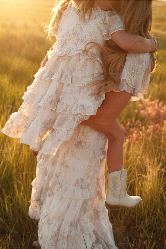 A woman in a ruffled white dress holds a young girl wearing the Ivy City Co Mini Winnie Dress in Bright White Wispy Wildflower and white cowboy boots in a sunlit field.