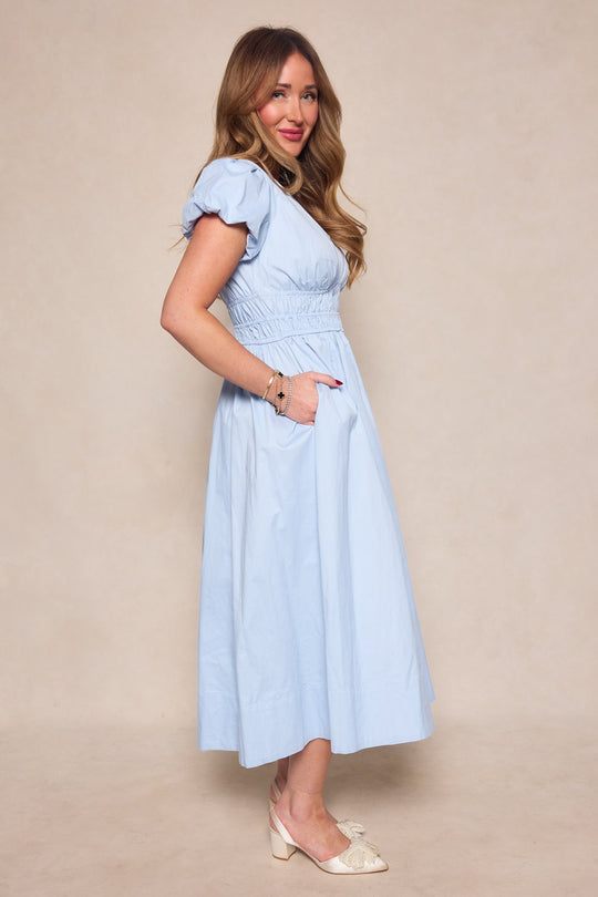 A woman with long hair stands sideways against a plain beige background, wearing the Ivy City Co Zella Dress in Sky Blue—a light blue, short-sleeve, midi dress with pockets—paired with light-colored heels.