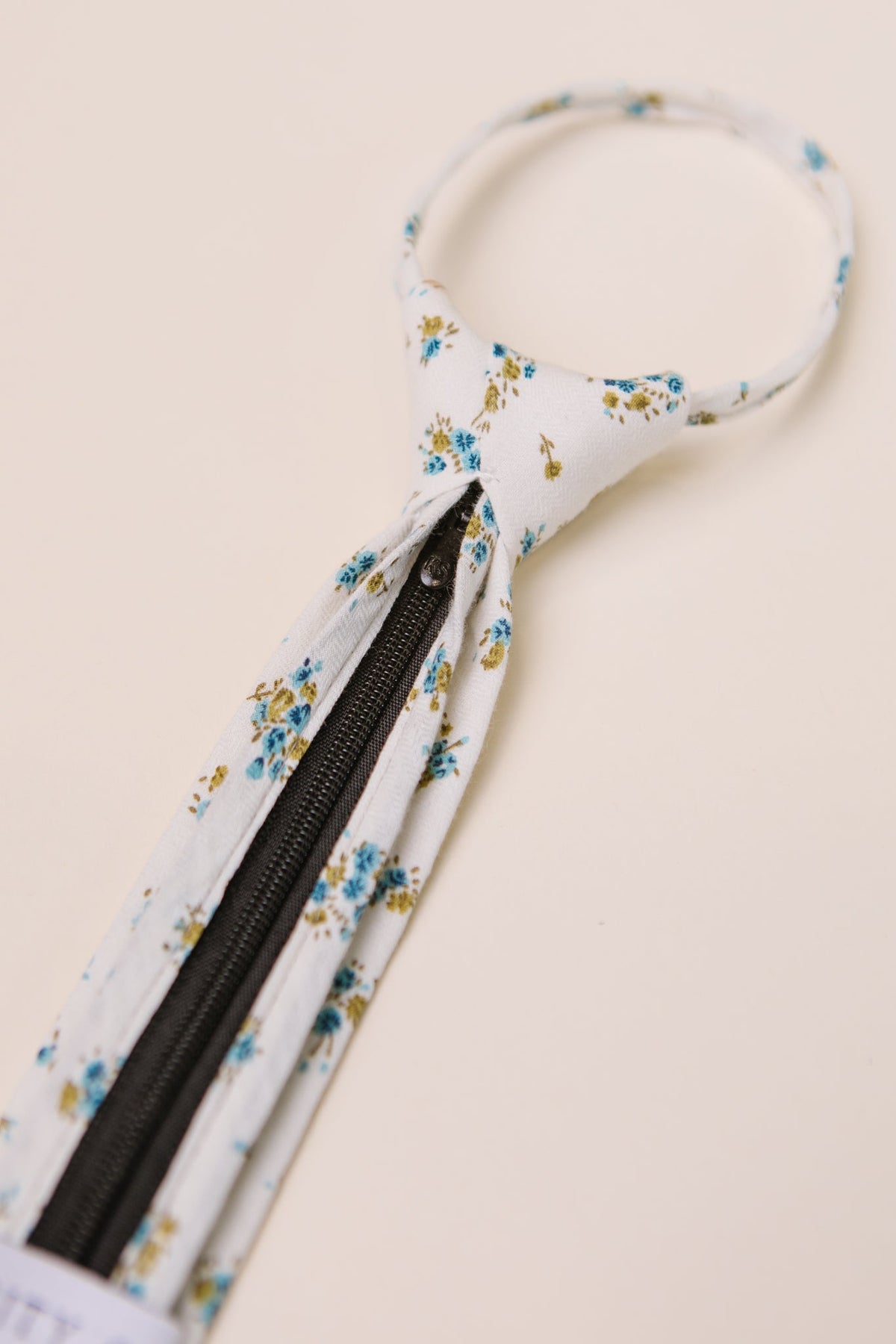 A white floral necktie adorned with blue and yellow blossoms and accented by a black center zipper is displayed on a light background—a stylish accessory that perfectly complements Ivy City Co’s Ties in Madeline Blue Floral for coordinated family matching.