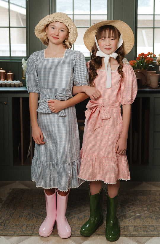 Two girls stand in a kitchen, smiling with arms linked. One wears Ivy City Cos Mini Hannah Dress in Blue Gingham and pink boots; the other wears a pink dress with green boots. Their wide-brimmed hats add vintage charm to the cheerful scene.
