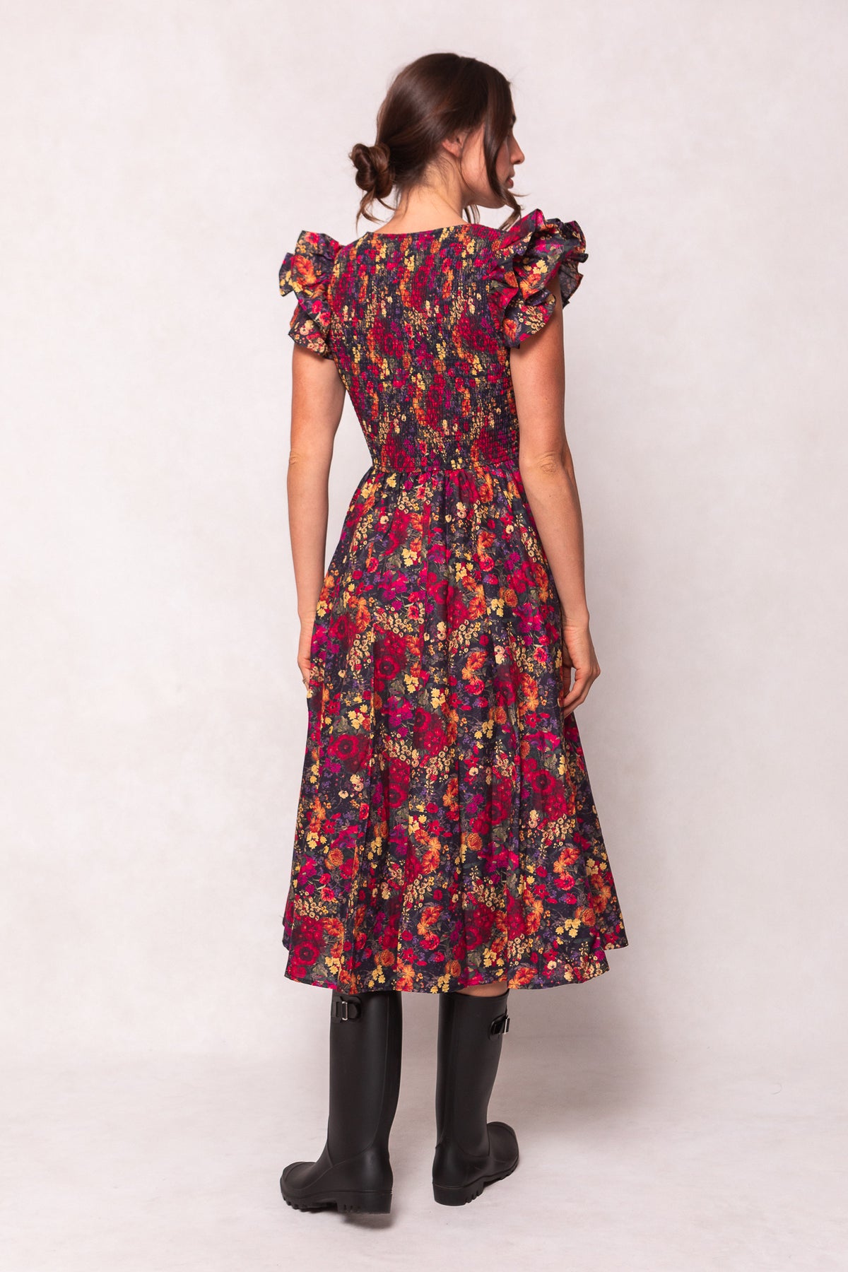 A woman faces away in the Ivy City Co Hattie Dress in Night Garden, a dark floral midi with ruffled shoulders and smocked bodice, paired with black knee-high boots. Her brown hair is in a low bun—an elegant maternity-friendly look.