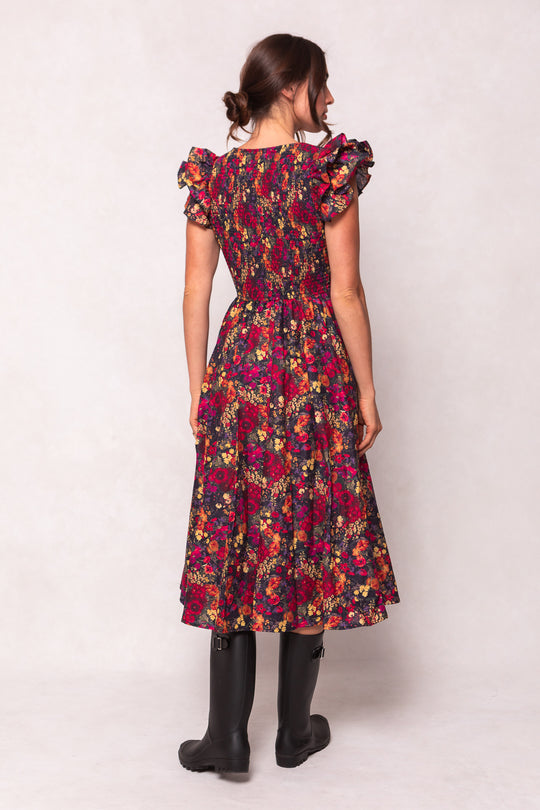 A woman faces away in the Ivy City Co Hattie Dress in Night Garden, a dark floral midi with ruffled shoulders and smocked bodice, paired with black knee-high boots. Her brown hair is in a low bun—an elegant maternity-friendly look.