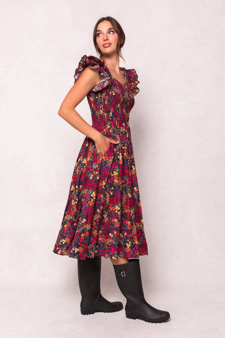 A woman in the Ivy City Co Hattie Dress in Night Garden stands against a light background, hands in pockets. She pairs the dark floral, maternity-friendly midi dress with black knee-high boots for an effortlessly chic look.