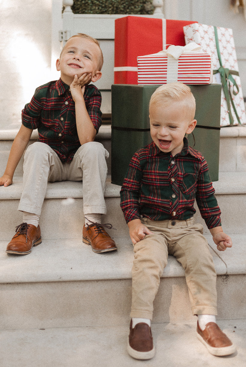 Two young boys in matching plaid shirts and khaki pants sit on steps in front of stacked wrapped gifts, smiling and laughing together—their cheerful outfits perfectly channel the festive spirit, just like Ivy City Co’s Baby Boys John Romper in Evergreen Plaid.