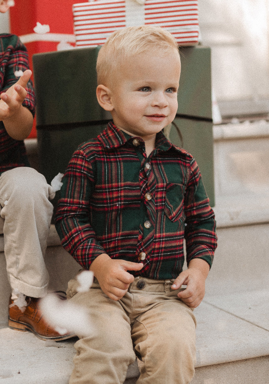 A young boy with blonde hair, wearing the Ivy City Co Baby Boys John Romper in Evergreen Plaid paired with beige pants, sits on steps in front of wrapped presents, smiling slightly.