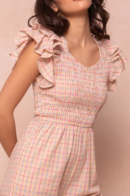 A woman wears the Ivy City Co Hattie Jumpsuit in Multicolor Gingham, featuring ruffled shoulders. Her dark wavy hair flows as she poses against a light beige background, with her face partially out of frame.