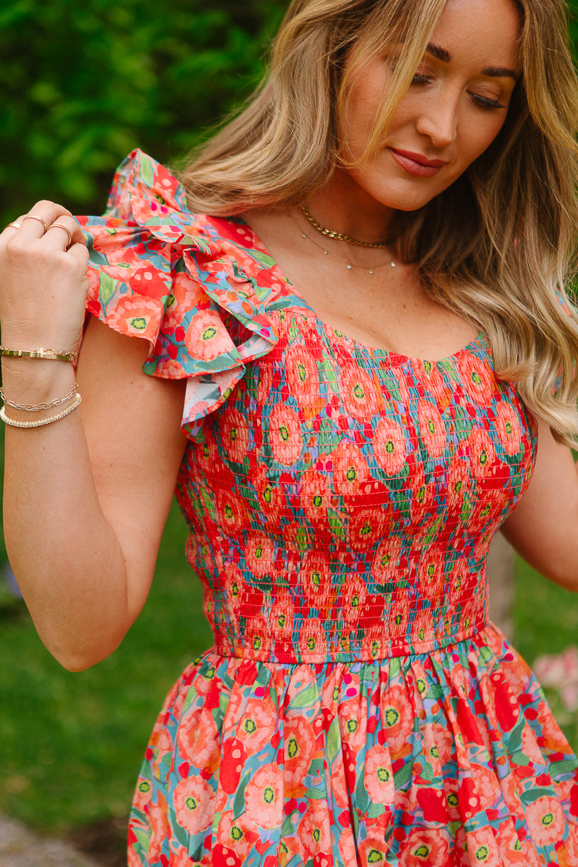 A woman with long, wavy blonde hair and gold jewelry stands outdoors in Ivy City Co’s Hattie Dress in Poppy, featuring cotton fabric and ruffled sleeves. She gently holds one sleeve, with blurred green foliage in the background.