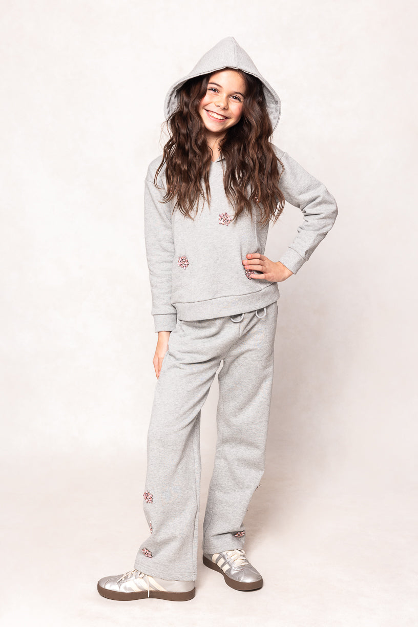 A smiling girl with long brown hair poses with one hand on her hip against a light background, wearing the Ivy City Co Mini Skylar Hoodie in Heather Gray, which features pink crystal flower accents, along with matching sweatpants.