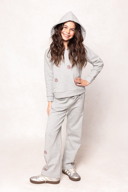 A smiling girl with long brown hair poses with one hand on her hip against a light background, wearing the Ivy City Co Mini Skylar Hoodie in Heather Gray, which features pink crystal flower accents, along with matching sweatpants.
