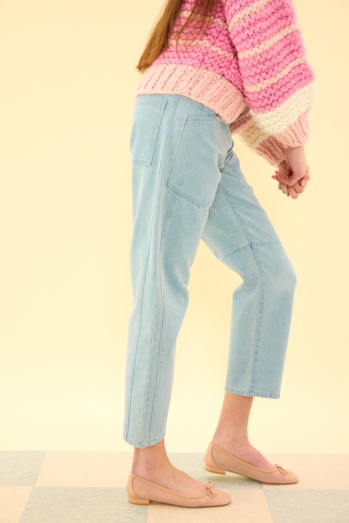A person wearing the Amanda Pants in Light Wash Denim by Ivy City Co, a pink and white striped knit sweater, and light pink flats stands on a checkered floor against a pale yellow background.