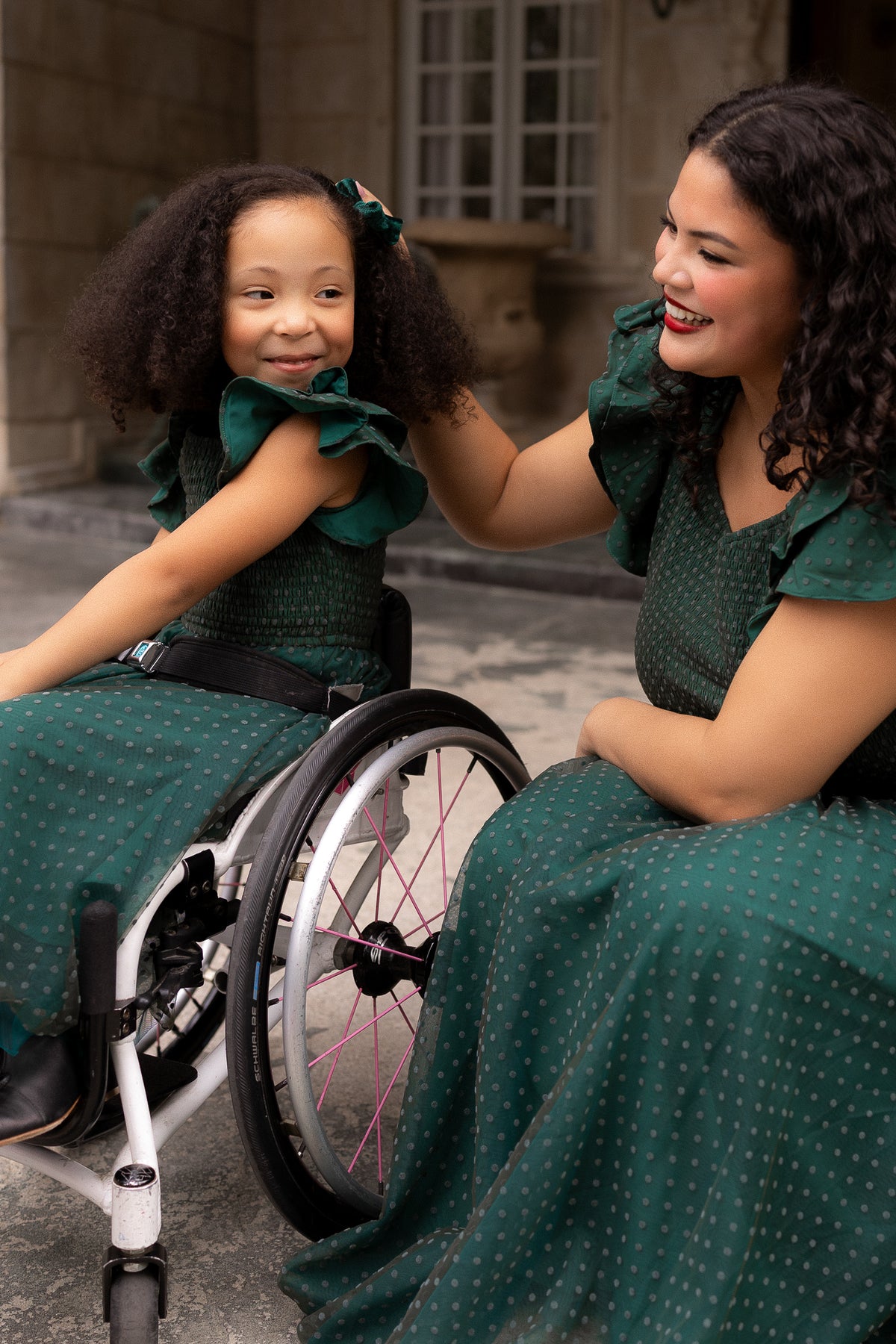 Two women in matching green dresses, little girl sitting in a wheelchair outdoors.
