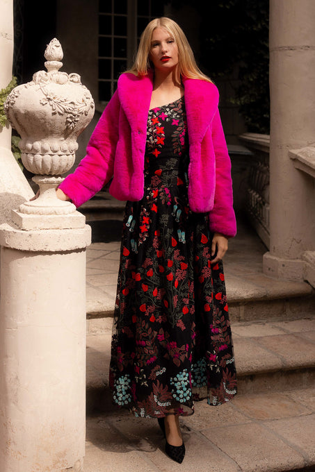 Woman in a floral dress and bright pink coat standing on stone steps.