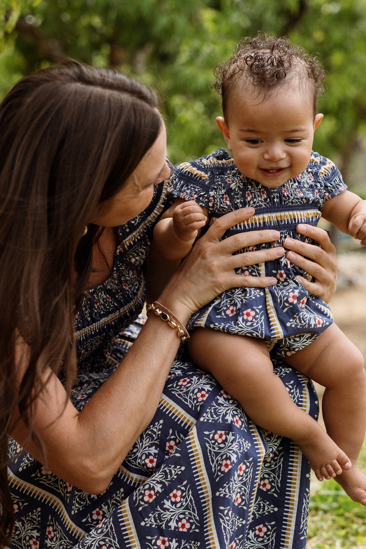 A woman holds a smiling baby outdoors amidst lush green foliage. Both are dressed in matching blue patterned outfits, with the baby wearing the Ivy City Co Baby Marlow Romper in Navy Fleur.