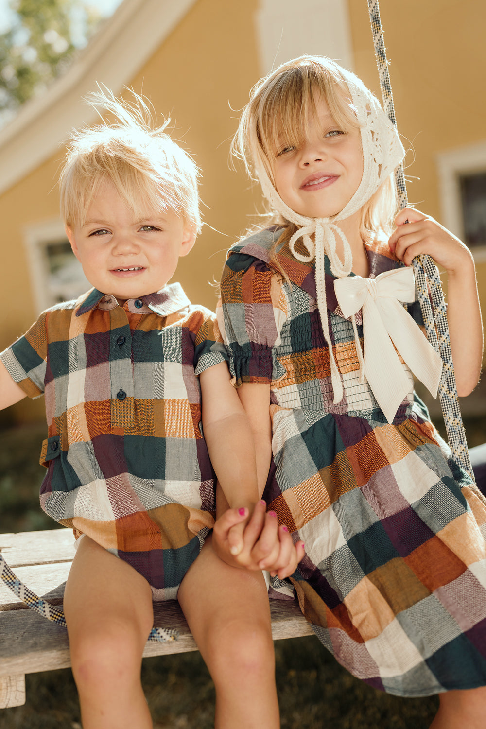 Two young children sit together on a swing outdoors, holding hands and smiling. They wear matching plaid outfits in warm tones; the girl’s Ivy City Co Mini Alice Dress in Maize Gingham features a smocked bodice and playful puff sleeves, complemented by a bonnet with a large bow. Sunlight bathes the scene, creating a bright and cheerful atmosphere.