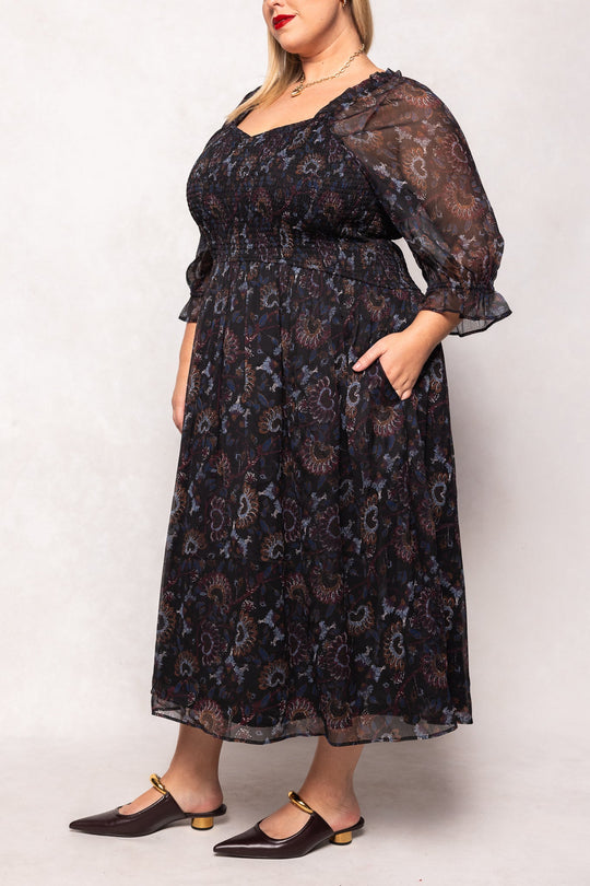 A woman stands against a light background wearing the Maddison Dress in Enchanted Floral by Ivy City Co—a dark, sheer midi dress adorned with a floral pattern, featuring puff sleeves and convenient pockets. Paired with brown low-heeled shoes, this elegant piece is maternity friendly, offering both comfort and style.
