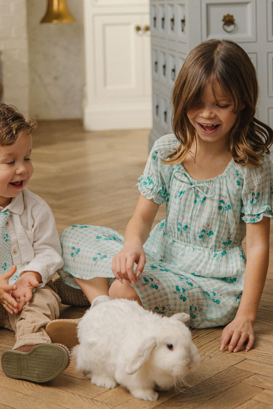 Two young children sit on a wooden floor, smiling as they watch a fluffy white rabbit. The girl—dressed in Ivy City Co’s Mini Jessie Dress in Bloom Gingham—gently pets the rabbit, while both children appear happy and engaged.