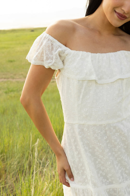 A woman wearing the Alaina Dress in Ivory by Ivy City Co—a white off-the-shoulder lace dress with a smocked bodice—stands in a grassy field, her face partially visible and one hand resting at her side.