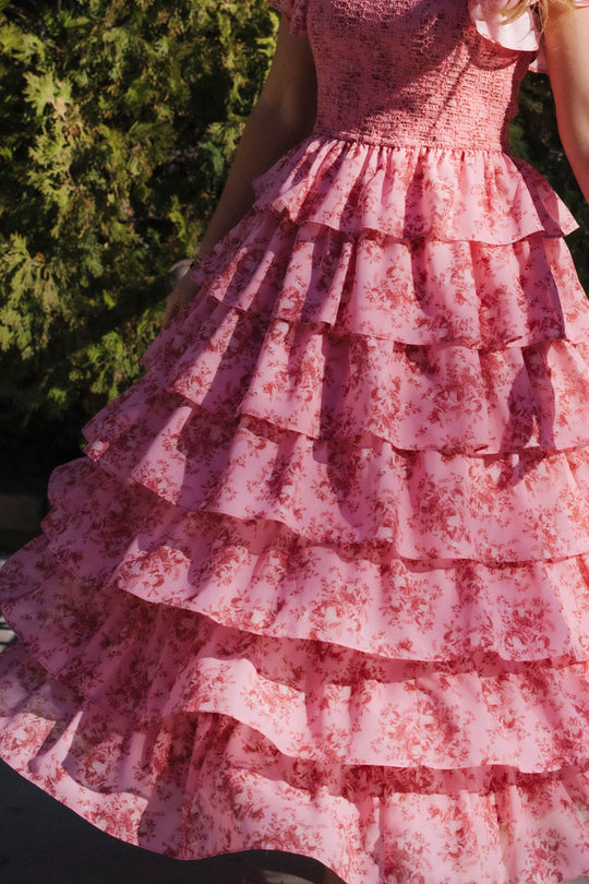 A person wearing the Ivy City Co Grace Dress in Desert Heritage Rose, featuring multiple ruffled layers and delicate floral patterns, stands outdoors near green bushes; only the lower half of this maternity-friendly dress is visible.