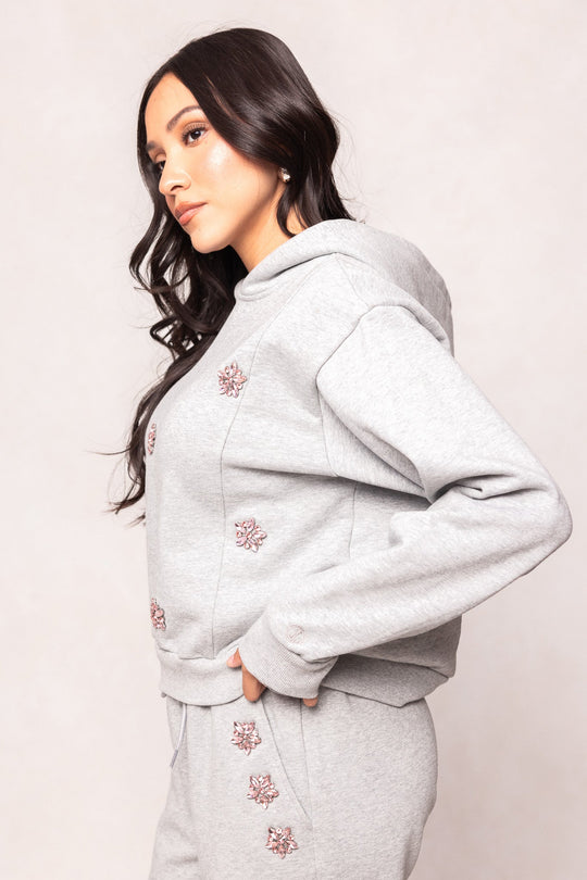 A woman with long dark hair stands sideways against a neutral background, wearing the Ivy City Co Skylar Hoodie in Heather Gray and matching sweatpants, featuring delicate pink flower embellishments.