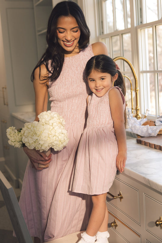 A woman, smiling and holding white flowers, stands beside a young girl in a bright, elegant kitchen with marble countertops and gold fixtures. Both are wearing Ivy City Co’s Karla Dress in Pink Striped Gingham.