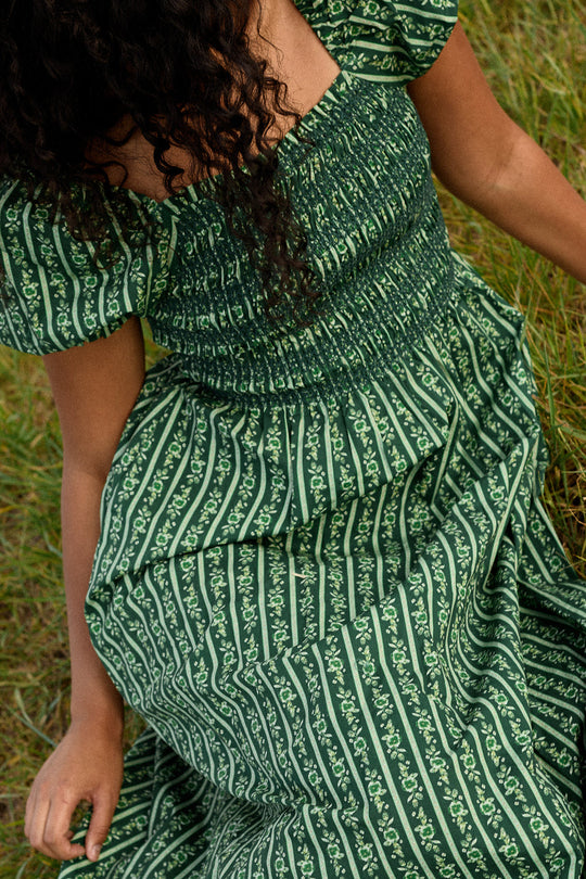 A person with curly hair sits on the grass, wearing the Ivy City Co Sunday Best Dress in Pine Felicity Floral, which features white vertical stripes and delicate floral patterns. This dress is both nursing and maternity friendly. Only their upper body and part of their face are visible.