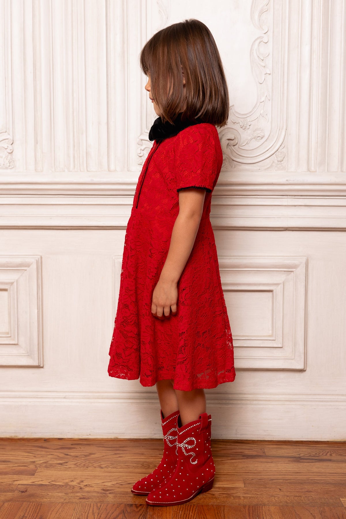 A young girl stands in profile against an ornate white wall with wooden floors, perfectly dressed for a special occasion in the Ivy City Co Mini Penny Dress in Red Lace and red studded boots.