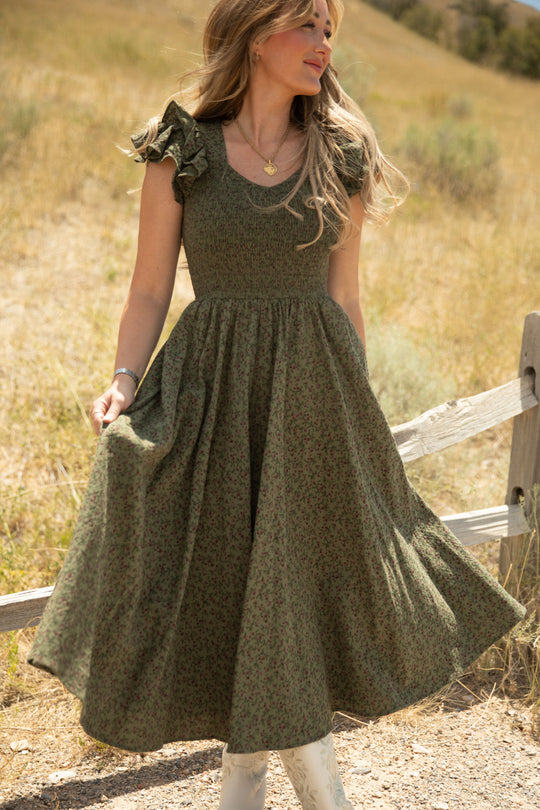A woman stands outdoors on a sunny day, holding the skirt of her Ivy City Co Hattie Dress in Woodland Floral. Maternity friendly and effortlessly stylish, the dress features a smocked bodice. She has wavy hair and wears white boots, with a wooden fence and grassy field in the background.