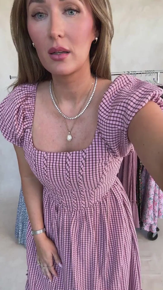 A woman stands indoors, looking at the camera while wearing the Monica Dress in Pink & Brown Gingham from Ivy City Co. The puff-sleeve midi dress features a square neckline and a charming gingham pattern. She accessorizes with a necklace and bracelet, her long hair styled down; behind her are clothing racks displaying more dresses.