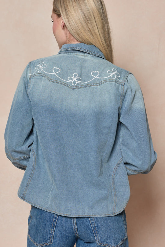 A person with long blonde hair wears the Denver Top in Lightwash Denim by Ivy City Co, featuring pearl snap buttons and white heart and swirl embroidery on the back, paired with blue jeans and standing against a beige background.