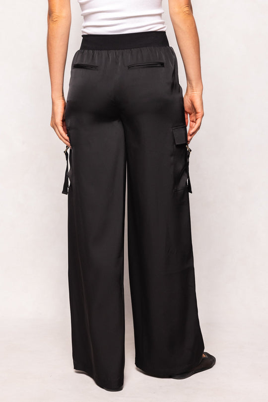 Someone is seen from the back standing against a light background, wearing the Shay Satin Pants in Black by Ivy City Co—loose-fitting cargo pants with an elastic waistband, wide legs, and side pockets.