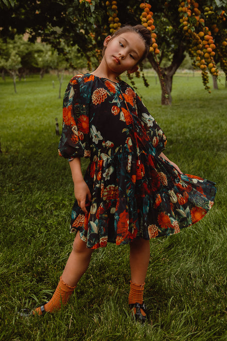 A young child poses outdoors on grass near a tree with orange fruit, wearing the Ivy City Co Mini Imogen Dress in Moody Floral and orange socks, tilting their head and holding out the skirt with one foot pointed.