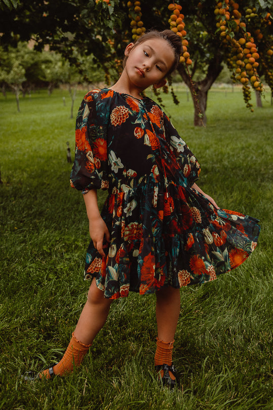 A young child poses outdoors on grass near a tree with orange fruit, wearing the Ivy City Co Mini Imogen Dress in Moody Floral and orange socks, tilting their head and holding out the skirt with one foot pointed.