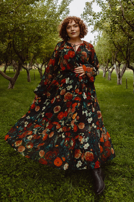 A woman with curly hair stands in a green orchard, confidently wearing the Ivy City Co Imogen Dress in Moody Floral—a stylish, maternity-friendly maxi—holding out her flowing patterned skirt.