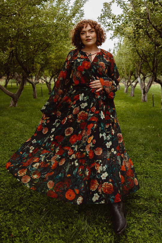 A woman with curly hair stands in a green orchard, confidently wearing the Ivy City Co Imogen Dress in Moody Floral—a stylish, maternity-friendly maxi—holding out her flowing patterned skirt.