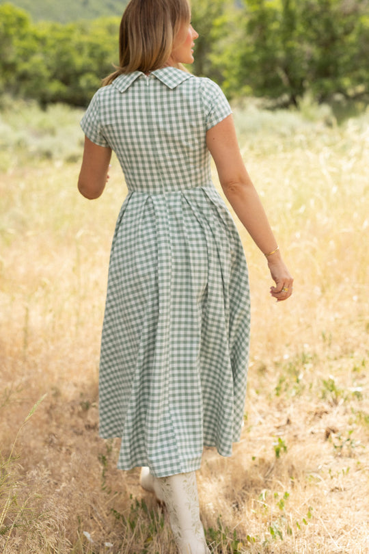 A woman wearing the Ivy City Co Amber Dress in Sage Gingham, featuring a pleated skirt, pairs it with white boots as she walks through a sunlit field surrounded by tall grass and trees.