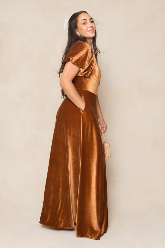 A woman stands sideways, smiling and holding a small boxy handbag, wearing the Ivy City Co Tessie Dress in Amber Velvet—a long, flowing dress with short puff sleeves. Her long hair is partially tied back with a white ribbon.