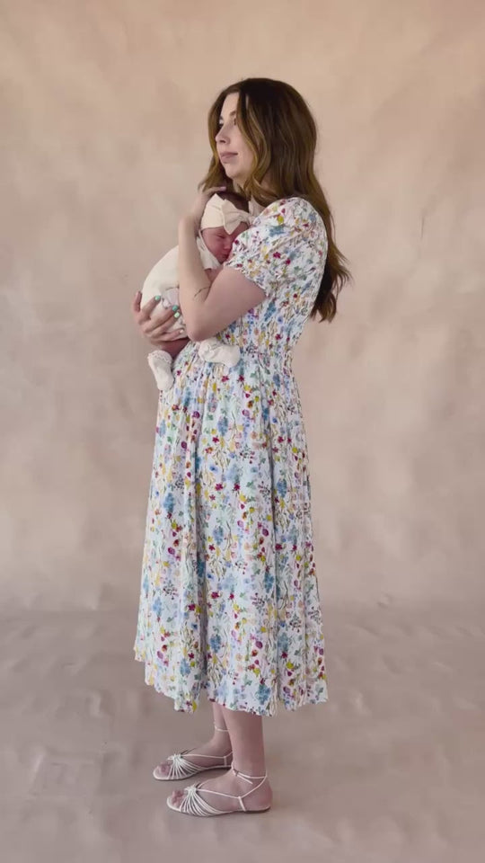 A woman wearing the Ivy City Co Amelie Dress in Floral Fields stands sideways, gently holding a baby wrapped in a white blanket against a neutral beige background.