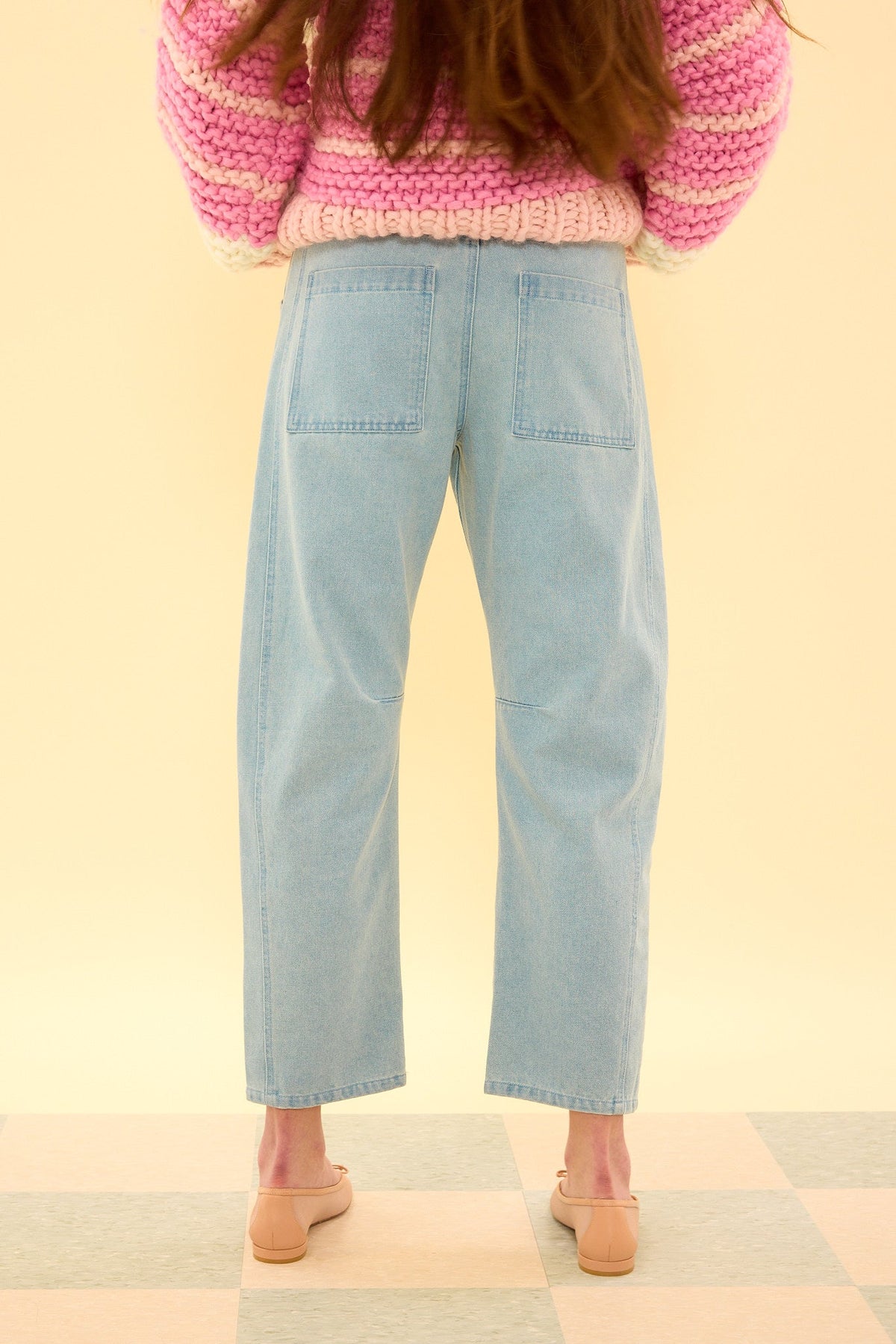 A person wearing the Ivy City Co Amanda Pants in Light Wash Denim, paired with a chunky pink and white sweater and beige shoes, stands on a tiled floor facing away from the camera against a cream background.