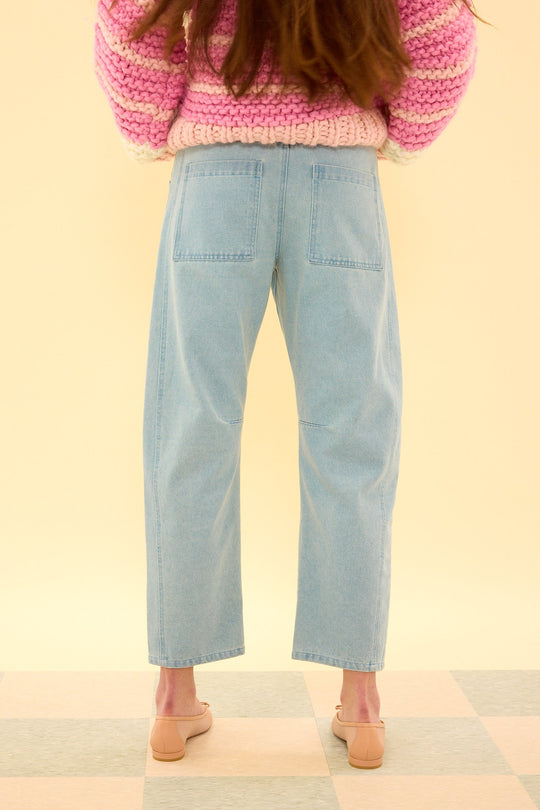 A person wearing the Ivy City Co Amanda Pants in Light Wash Denim, paired with a chunky pink and white sweater and beige shoes, stands on a tiled floor facing away from the camera against a cream background.