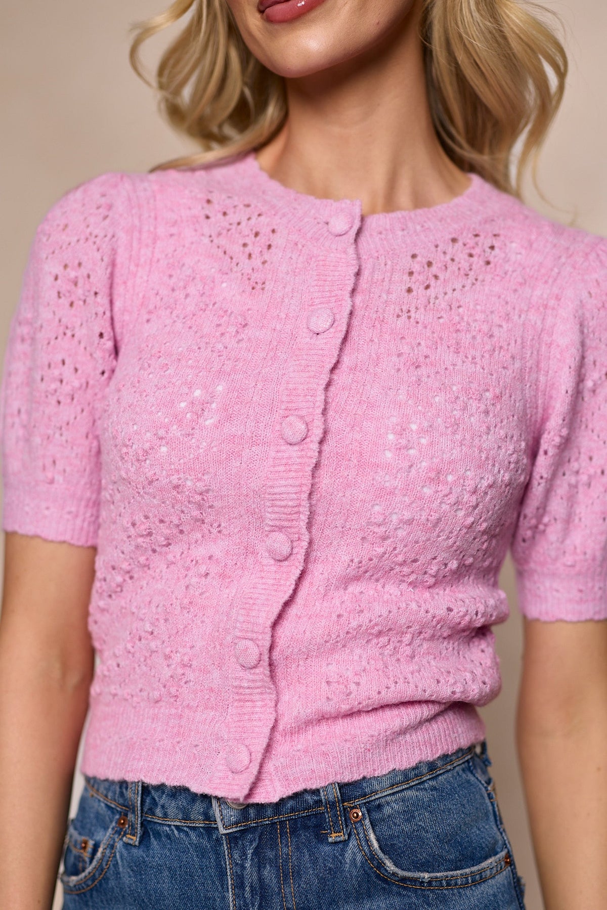 A woman wearing the Michaela Cardigan in Pink by Ivy City Co—a light pink, short-sleeve, fitted button-front knitted sweater with eyelet details—paired with high-waisted blue jeans.