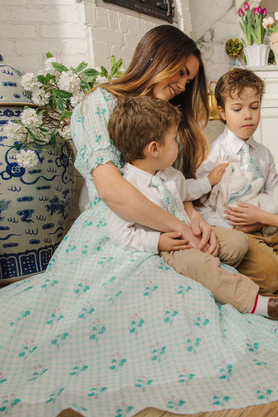 A woman wearing the Jessie Dress in Bloom Gingham by Ivy City Co, a maternity-friendly pastel floral design, sits indoors with two young boys—one holding a white bunny—beside a large vase of flowers and a tulip-adorned mantel.