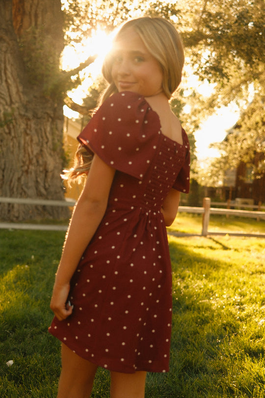 A girl stands on the grass outdoors, looking back over her shoulder in the Ivy City Co Teen Blakely Dress in Cranberry Dainty Dot. Sunlight filters through the trees behind her, casting a warm, golden glow that highlights the dress’s delicate petal sleeves and charming white dots on its midi-length silhouette.