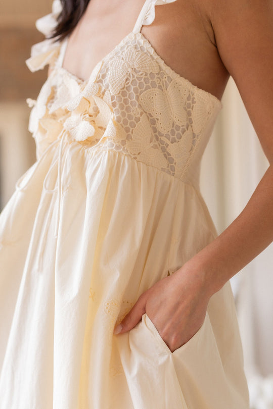 A person wearing the Ivy City Co Gaia Butterfly Dress in Ivory—a cream-colored maxi dress featuring lace and butterfly embroidery, ruffled straps, and a side pocket with their hand in it. The dress is maternity friendly, and only the upper body is visible.