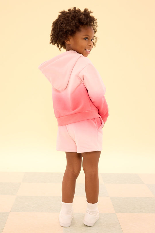 A young child with curly hair, wearing the Ivy City Co Mini Ash Hoodie in Pink Ombre and matching pink ombre sweatshorts, stands on a checkered floor and smiles over their shoulder against a light yellow background.